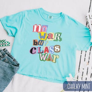 May include: A light blue t-shirt with the text "no war but class war" in a colorful, cut-out style. The shirt is a crew neck and has short sleeves. The color is "chalky mint".