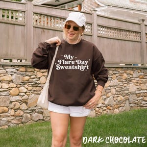 May include: A dark brown long-sleeve sweatshirt featuring the white text "My Flare Day Sweatshirt" with small star accents. The garment is paired with a white baseball cap, sunglasses, and a white shoulder bag with a strap.