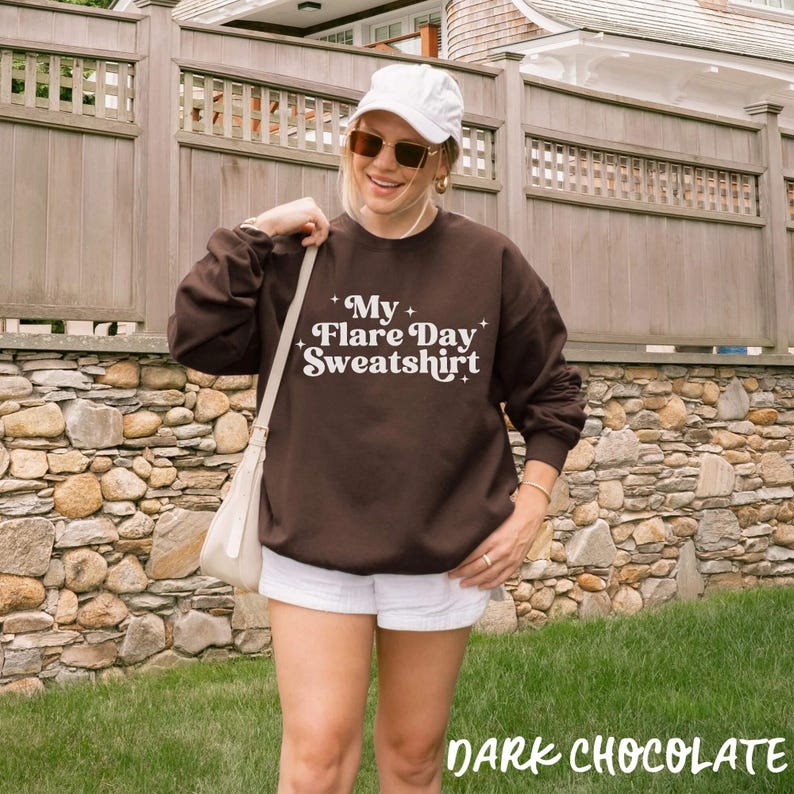 May include: A dark brown long-sleeve sweatshirt featuring the white text "My Flare Day Sweatshirt" with small star accents. The garment is paired with a white baseball cap, sunglasses, and a white shoulder bag with a strap.