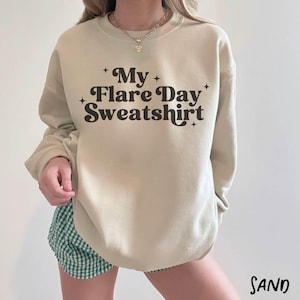 May include: A sand-colored sweatshirt with the text "My Flare Day Sweatshirt" in a black, retro font. The sweatshirt has a crew neck and long sleeves. The word "SAND" is printed in a sans-serif font at the bottom right.