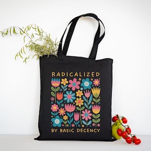 May include: Black tote bag with a colorful floral design and the text "RADICALIZED BY BASIC DECENCY" in gold. The flowers are in shades of pink, blue, yellow, and orange. The bag has black handles and is on a white background.