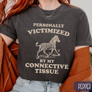May include: Dark gray t-shirt with the text "PERSONALLY VICTIMIZED BY MY CONNECTIVE TISSUE" above and below a zebra illustration. The shirt is a crew neck and has short sleeves.