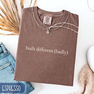 Peut inclure: T-shirt couleur expresso avec l'inscription "built different (badly)". Le t-shirt est plié et orné d'un collier argenté avec un pendentif circulaire. Un jean bleu et une chaussure blanche sont visibles.