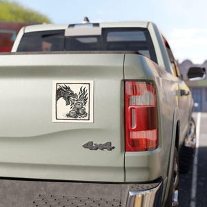 May include: A square sticker featuring a black and white illustration of a dragon breathing fire onto a pile of cubes. The sticker is affixed to the back of a light green pickup truck.