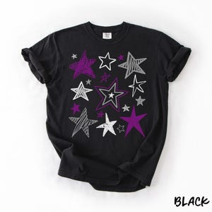 May include: Black t-shirt with a star design. The design features various star shapes in white, purple, and gray. The word "BLACK" is printed in white at the bottom right corner of the shirt.