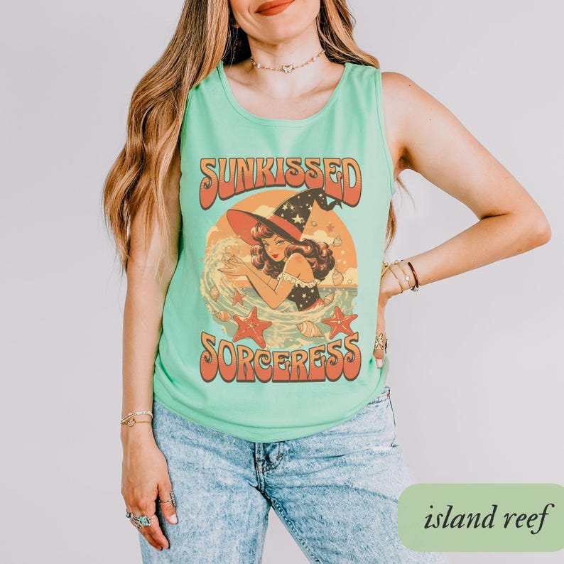 May include: A light green tank top with the words "SUNKISSED SORCERESS" in orange lettering. The graphic features a woman in a witch hat and a red and orange sunset background. The tank top is paired with blue jeans.