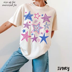 Peut inclure: T-shirt ivoire avec un motif d'étoiles bleues, roses et violettes. Le texte "size up for oversized fit" est au-dessus de l'épaule. Le mot "IVORY" est imprimé en bas à droite. Le t-shirt est de style décontracté.