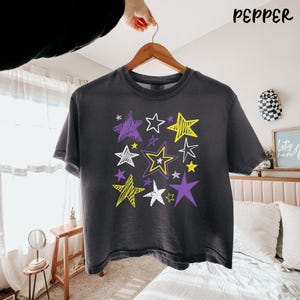 May include: A charcoal gray short-sleeve t-shirt featuring a graphic print of various stars in yellow, purple, and white. The stars are rendered in a hand-drawn, scribbled style with different sizes and outlines scattered across the center chest.