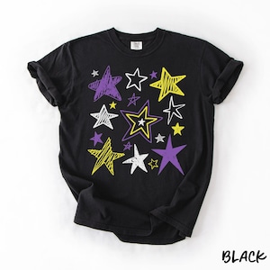 May include: A black short-sleeve t-shirt featuring a graphic print of multiple stars in various sizes and styles. The stars are rendered in white, yellow, and purple with scribbled, outlined, and solid fill designs scattered across the front.