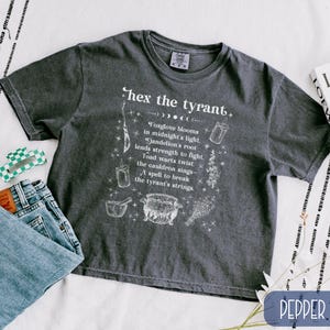 May include: Dark grey t-shirt with the text "hex the tyrant" and a spell graphic. The shirt also features a list of spell ingredients. A pair of blue jeans and a green and white hair clip are visible.
