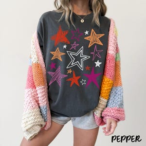 May include: Dark gray sweatshirt with colorful star graphics in orange, red, pink, and white. A multi-colored, patchwork cardigan is worn over the sweatshirt. The word "PEPPER" is printed in the bottom right corner.