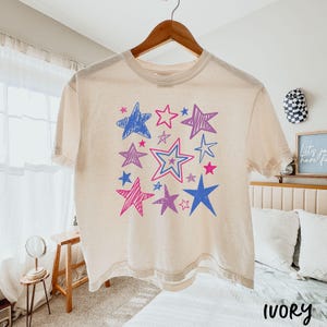 May include: Ivory-colored t-shirt with a star design in shades of blue, pink, and purple. The stars vary in size and style, creating a playful pattern. The shirt is hanging on a wooden hanger.