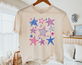 May include: Ivory-colored t-shirt with a star design in shades of blue, pink, and purple. The stars vary in size and style, creating a playful pattern. The shirt is hanging on a wooden hanger.
