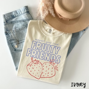 May include: Cream-coloured t-shirt with the text "support your local FRUITY FRIENDS" in blue and red, featuring two strawberry illustrations. The shirt is paired with light blue jeans, a beige hat, and a necklace.