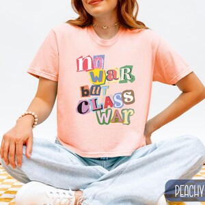 May include: A peach-colored t-shirt with the text "no war but class war" in a colorful, cut-out style. The shirt has short sleeves and a crew neck. The model is wearing light blue jeans and white sneakers.
