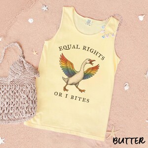 Puede incluir: Una camiseta sin mangas de color amarillo claro con el texto "EQUAL RIGHTS OR I BITES" y un ganso con alas de arco&iacute;ris. A la izquierda hay un bolso de ganchillo beige. La palabra "BUTTER" est&aacute; en la parte inferior derecha.