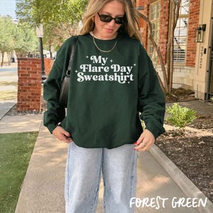 May include: A forest green sweatshirt with the text "My Flare Day Sweatshirt" in white. The sweatshirt is paired with light wash jeans. The words "Forest Green" are at the bottom of the image.