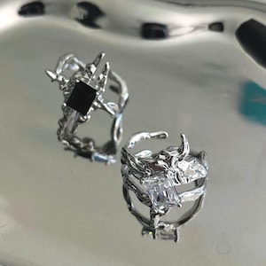 Può includere: Due anelli d'argento con design organici intricati. Un anello presenta una pietra quadrata nera, l'altro una pietra rettangolare trasparente. Gli anelli sono esposti su una superficie argentata riflettente.