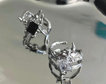 Könnte beinhalten: Zwei silberne Ringe mit aufwendigen, organischen Designs. Ein Ring hat einen schwarzen quadratischen Stein, der andere einen klaren rechteckigen Stein. Die Ringe werden auf einer reflektierenden silbernen Oberfläche gezeigt.