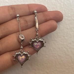 May include: A pair of silver earrings with a pink heart charm. The heart charm is surrounded by a silver crown design. The earrings are hanging from a silver chain.