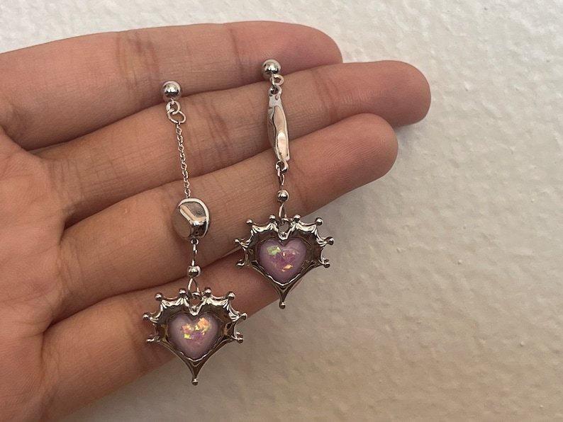 May include: A pair of silver earrings with a pink heart charm. The heart charm is surrounded by a silver crown design. The earrings are hanging from a silver chain.