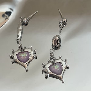 May include: A pair of silver dangle earrings with a heart-shaped charm featuring a pink iridescent stone. The heart is surrounded by a crown-like design.