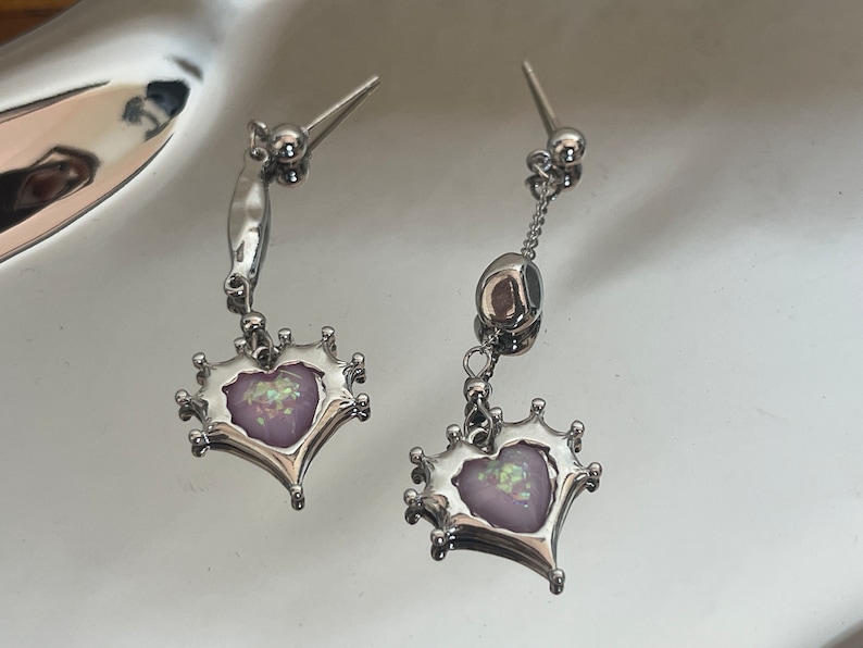 May include: A pair of silver dangle earrings with a heart-shaped charm featuring a pink iridescent stone. The heart is surrounded by a crown-like design.