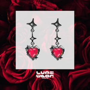May include: A pair of black dangle earrings featuring a red heart-shaped crystal and star accents. The earrings are displayed against a white backdrop.