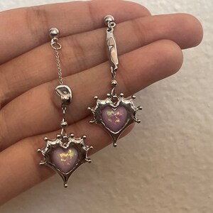 May include: A pair of silver earrings with a dangling heart-shaped charm. The hearts are adorned with a pink and iridescent stone.