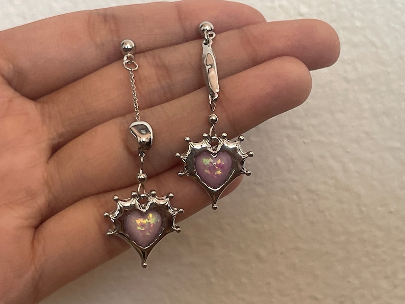 May include: A pair of silver earrings with a dangling heart-shaped charm. The hearts are adorned with a pink and iridescent stone.