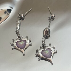May include: A pair of silver earrings with a dangling heart-shaped charm. The heart is decorated with a pink and iridescent design.