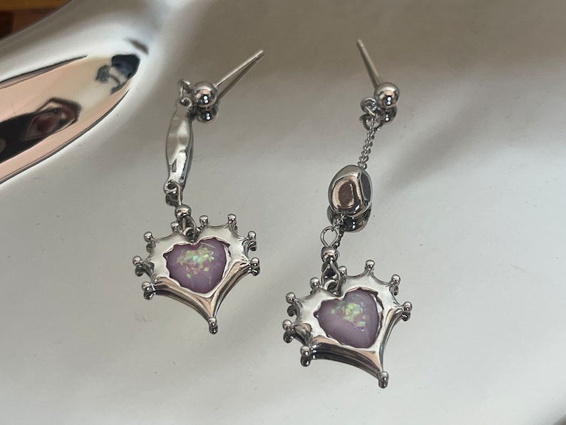 May include: A pair of silver earrings with a dangling heart-shaped charm. The heart is decorated with a pink and iridescent design.