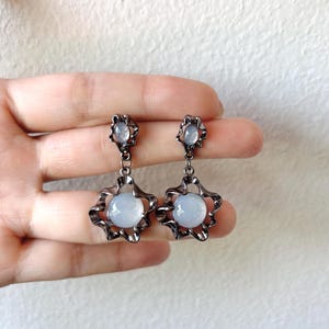 May include: A pair of silver dangle earrings with a white stone in the centre of each earring. The earrings have a floral design.