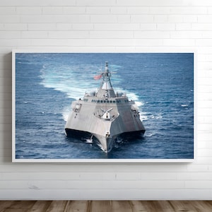 Puede incluir: Impresión enmarcada de un buque de guerra gris navegando por aguas azules, dejando una estela blanca. El barco, con una bandera americana, se presenta sobre una pared de ladrillos blancos.