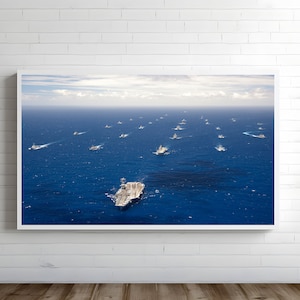 May include: An aerial view of a large naval fleet sailing on a deep blue ocean. The image shows numerous warships, including aircraft carriers and support vessels, spread across the water under a partly cloudy sky. The ships are arranged in formation.