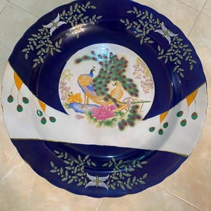 May include: A round ceramic plate featuring a deep blue border with green leaf patterns and white accents. The centre displays a colourful illustration of a peacock with a fanned tail, a smaller bird, and pink flowers on a white background.