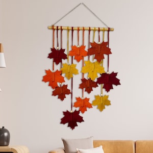 Pode incluir: Uma tapeçaria de parede com uma coleção de folhas de bordo de feltro coloridas em tons de vermelho, laranja e amarelo. As folhas estão suspensas em uma haste de madeira e estão dispostas em um padrão em cascata.