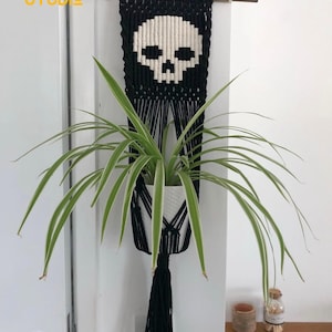 Op de afbeelding: Een zwarte macramé plantenhouder met een wit schedelontwerp. De plantenhouder is opgehangen aan een houten deuvel en heeft een witte keramische pot met een groene plant. De tekst "TINY STUDIO" staat in de linkerbovenhoek.