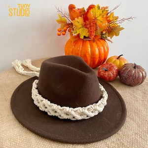 May include: A brown fedora hat with a white macrame band and tassels. The hat is sitting on a hessian surface in front of a pumpkin and autumn foliage.