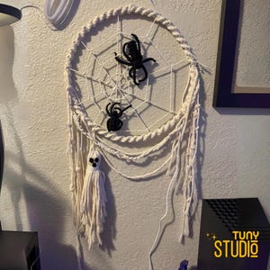 May include: A cream-colored macrame dream catcher with a spiderweb design. Two black spiders are attached to the web. A white ghost-shaped tassel hangs below. The dream catcher is a Halloween decoration.