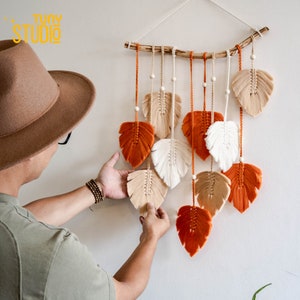 May include: A wall hanging made of macrame with orange, white, and beige feathers. The feathers are attached to a wooden branch and hang from the ceiling.