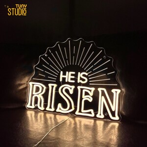 May include: White neon sign with the words "He is Risen" in a black background. The sign has a sunburst design behind the words.
