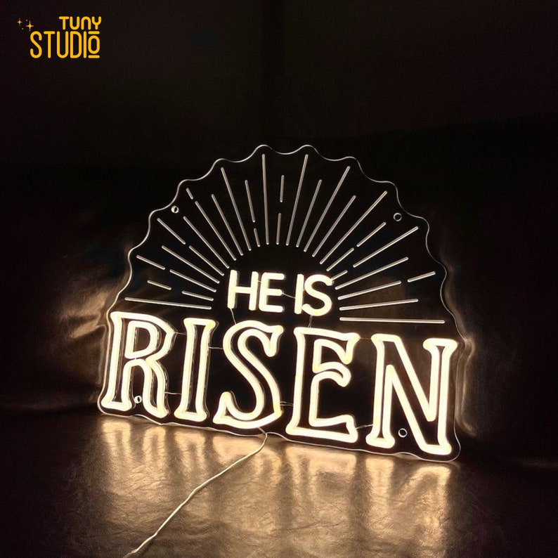 May include: White neon sign with the words "He is Risen" in a black background. The sign has a sunburst design behind the words.