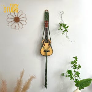 May include: A ukulele suspended in a green macrame plant hanger against a white wall. The ukulele has a natural wood finish. A daisy-shaped wall decoration and greenery are also visible.