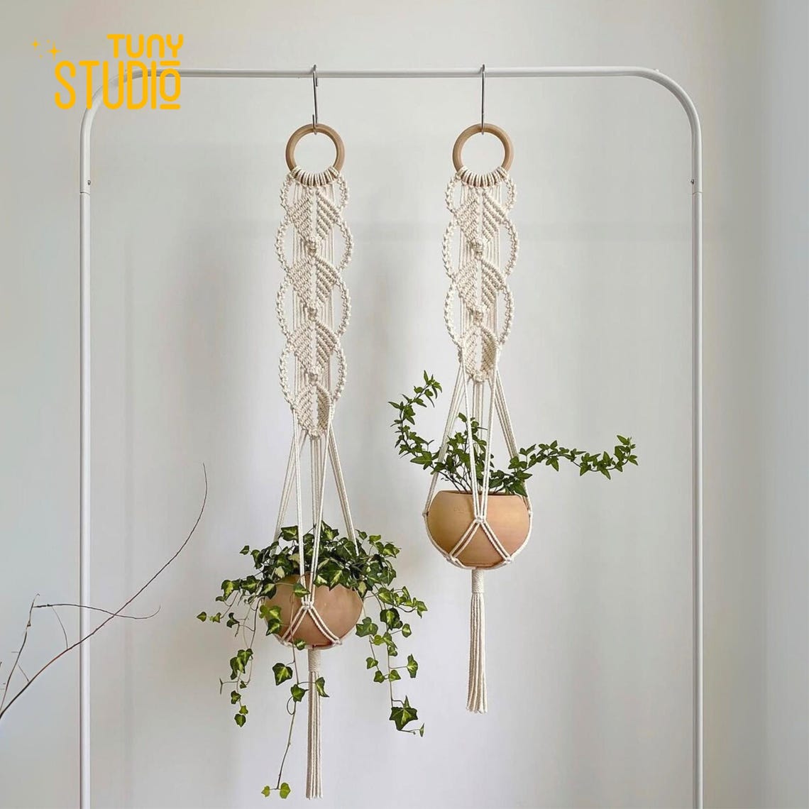 May include: Two macrame plant hangers with beige rope and wooden rings. Each hanger suspends a round, tan-colored pot filled with green plants. The hangers are suspended from a white metal bar against a white background. The word 'STUDIO' is visible in yellow.