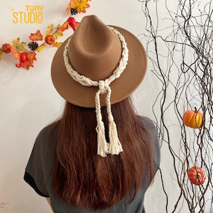 Pode incluir: Um chapéu fedora de feltro marrom com uma faixa de corda trançada branca e borlas. O chapéu é usado por uma pessoa com cabelo longo e castanho.