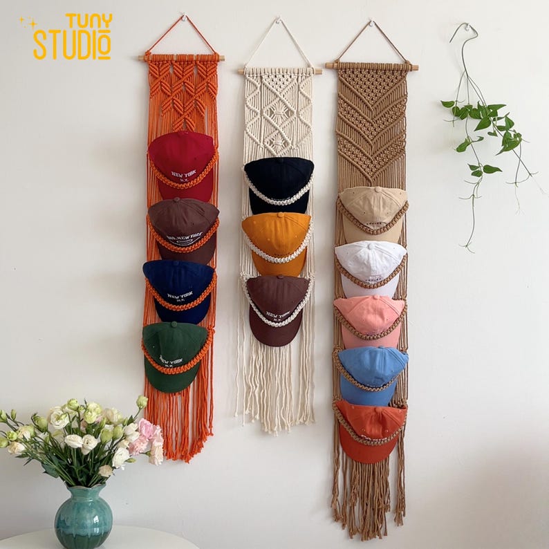 May include: Three macrame hat organizers in orange, cream, and brown hang on a white wall. Each organizer holds several baseball caps in various colors, including red, brown, navy, green, black, yellow, white, pink, blue, and orange.