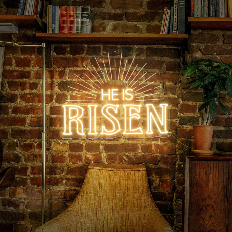 May include: A neon sign with a white outline and yellow glow, reading "He is Risen" with a sunburst design behind the text. The sign is mounted on a brick wall.