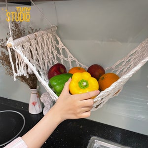May include: White cotton rope hammock hanging from a kitchen cabinet, filled with fresh produce, including red apples, yellow and green bell peppers, and oranges.