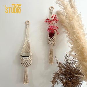 May include: Two macrame plant hangers with wooden rings, one holding a pot with red flowers. The hangers are made of cream-coloured woven cotton. The logo "TUNY STUDIO" is in yellow letters in the upper left corner.